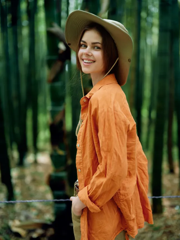 Woman smile hat forest bamboo portrait of young female in orange shirt, casual outdoor portrait with hands in pockets, happy traveler leaning on fence in green nature with relaxed expression.