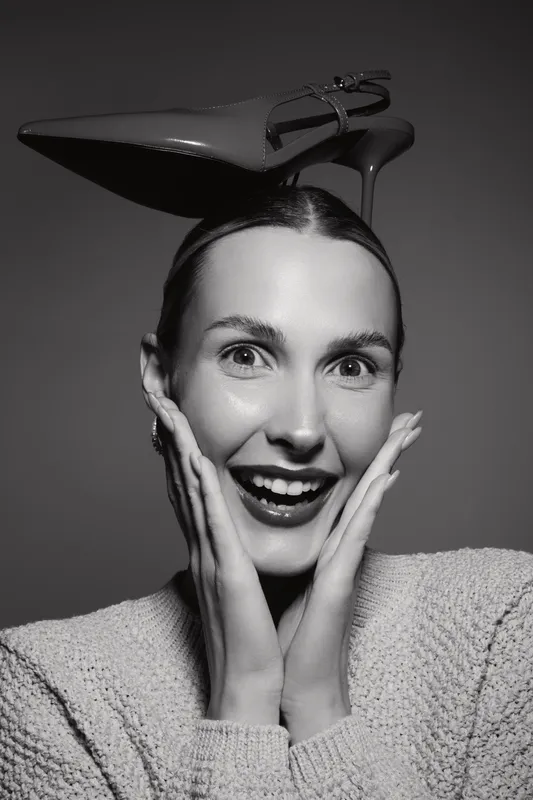 Happy Caucasian woman with light skin and sleek hair holds hands near face with excitement, balancing a high heel shoe on her head in studio. Concept of fun and fashion.