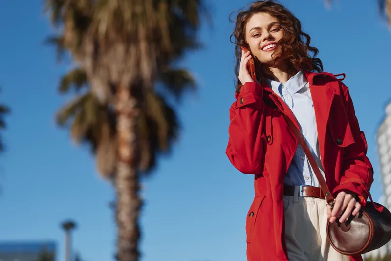 Cheerful woman outdoors wearing a red coat, blue shirt, and beige pants, sunlit moment with wind in hair, casual pose against palm trees and clear blue sky