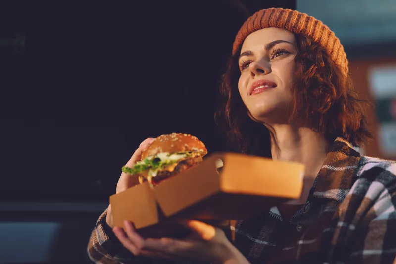 Woman holding burger in takeaway box, smiling in beanie and plaid shirt at night with golden hour glow, candid lifestyle authenticity and mindful living captured in emotional storytelling.