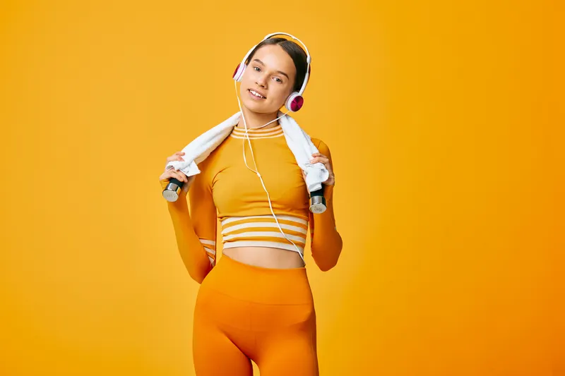 Woman in bright orange athletic outfit with headphones, holding dumbbells and a towel draped over shoulders, performing a high-energy workout against a vivid orange backdrop.