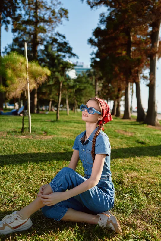 young woman sitting on grass in park wearing denim outfit and sunglasses on sunny day with trees background relaxing outdoors