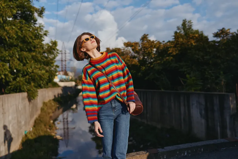 Young woman in a colorful striped sweater and sunglasses, short hair, wearing jeans, leaning back on an urban bridge under sunny sky. Casual outfit, travel lifestyle mood.