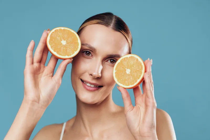A smiling young woman holds two orange halves near her face against a blue background. Concept for healthy lifestyle, natural beauty, and fresh fruit consumption.