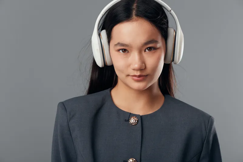 Young woman with long black hair wearing white headphones and gray jacket, looking at camera with neutral expression on plain gray background in studio portrait style.
