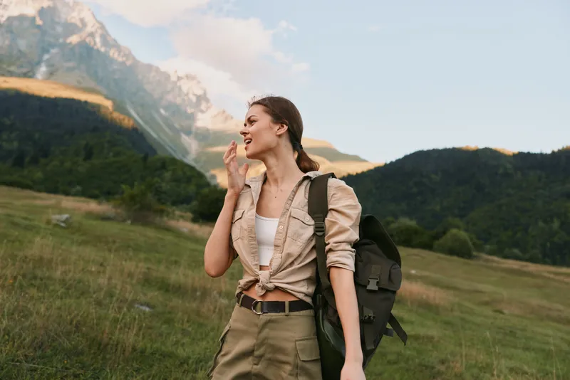 Young woman with a backpack enjoys a mountain hike while smiling and looking to the side in a green meadow under a partly cloudy sky.
