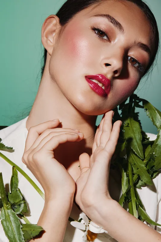 Fashion creative beauty portrait of woman with glossy pink makeup and bold lips posing with hands near her face holding green leaves on mint background. Concept for modern style.