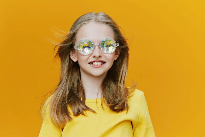 Happy girl with long hair wearing bright yellow shirt and colorful sunglasses against vivid yellow background, embodying joy and summer vibes