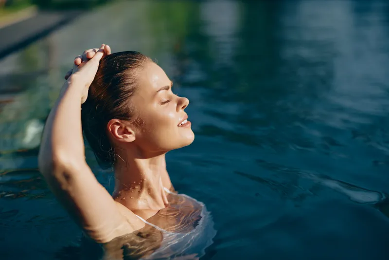 Relaxed woman enjoys a sunny outdoor swim, eyes closed, head tilted back, basking in warm light as she rises from clear water during a peaceful summer moment.