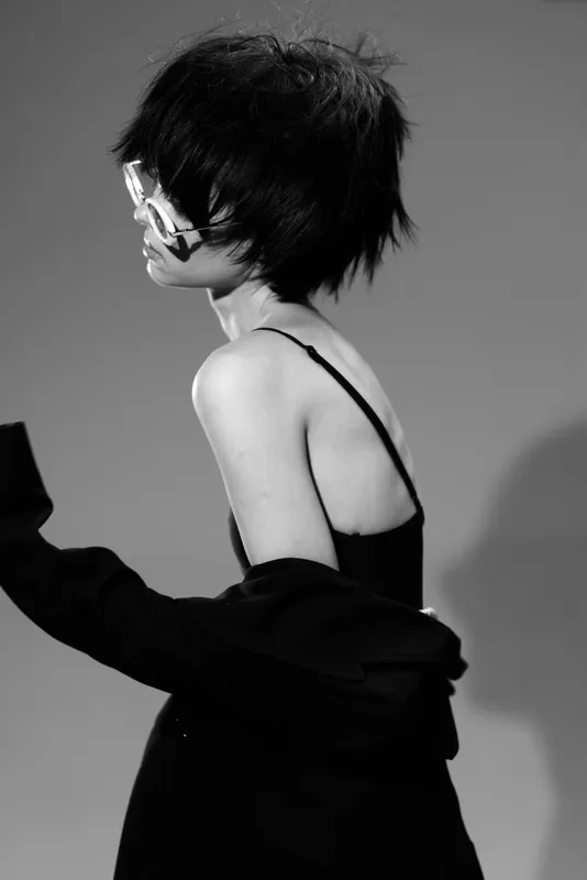 Black and white portrait of a thoughtful woman with glasses in stylish clothing posing indoors with dramatic lighting and shadows.
