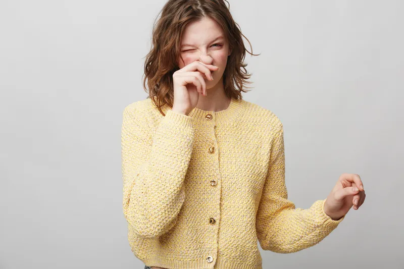 Young woman in yellow knitted sweater laughs and covers mouth with hand, standing against plain grey background. Casual style, happiness, joy, and candid moment concept.