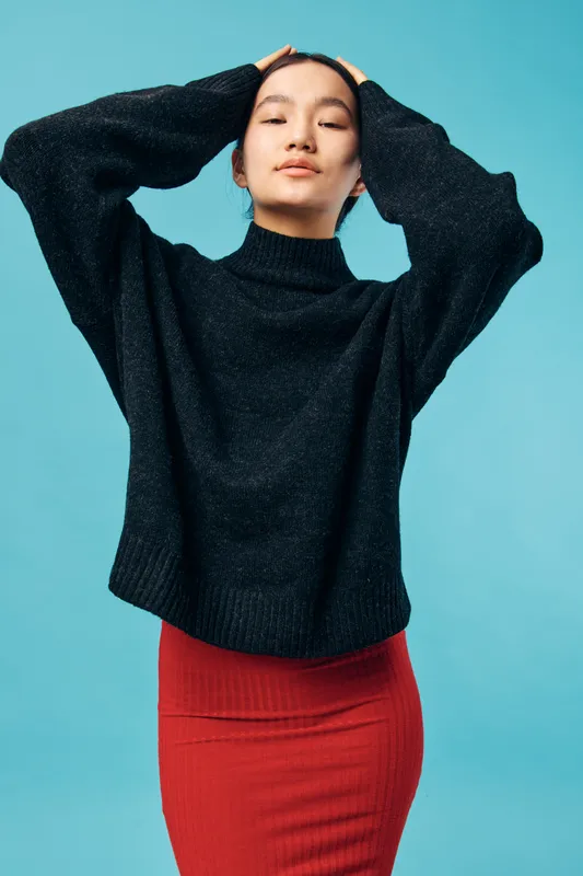 Young woman wearing black turtleneck sweater and red skirt posing with hands on head against blue background, casual fashion portrait of confident female model in studio.