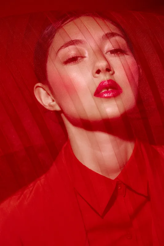 Stylish woman with glossy red lips looks through sheer red fabric with elegant intensity, wearing matching vibrant clothing and makeup. Creative fashion beauty concept illuminated by soft light.