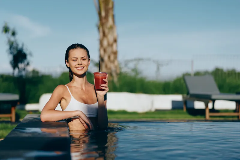 Happy woman relaxes in a poolside setting, holding a red drink, smiling at the camera, sunny outdoor scene with palm tree and lounge chairs, lifestyle moment of refreshment and leisure.