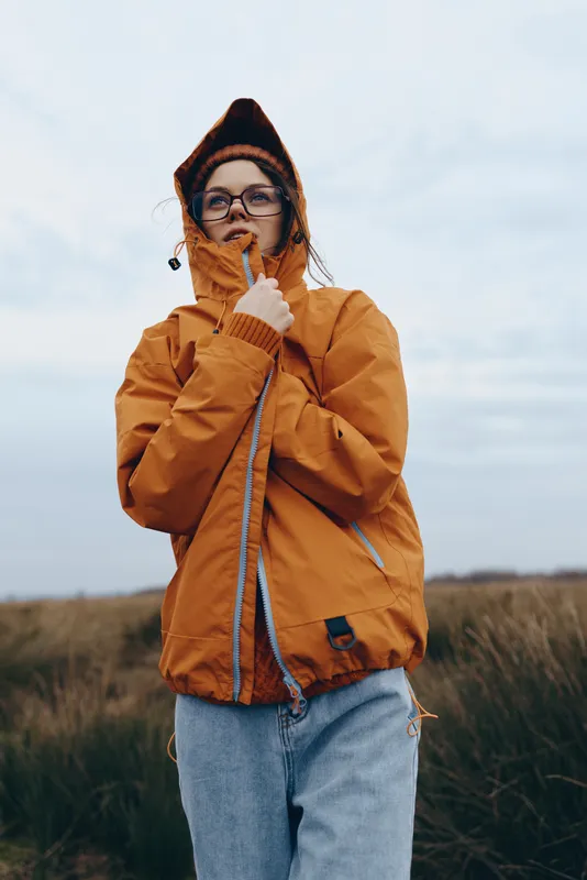 Woman outdoors wearing orange jacket with hood, glasses, covering mouth with hand against cold wind, blowing nose, under cloudy sky and grassy field, lifestyle seasonal illness concept
