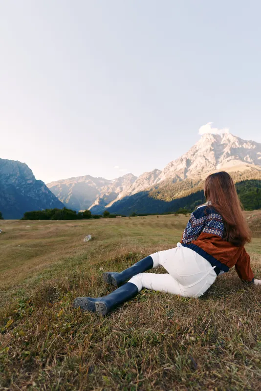Woman sitting on grassy meadow facing mountains and nature landscape in autumn, wearing cozy sweater, jacket and boots, relaxing outdoors with long hair and peaceful view