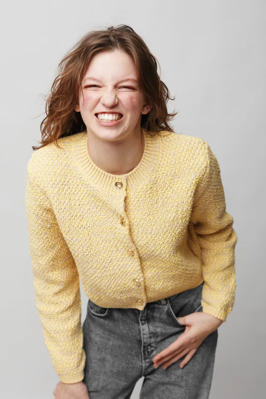Young woman in yellow sweater and grey jeans smiles with eyes closed and clenched teeth. Casual style, studio portrait on light background. Concept of joy, emotion, and fashion.