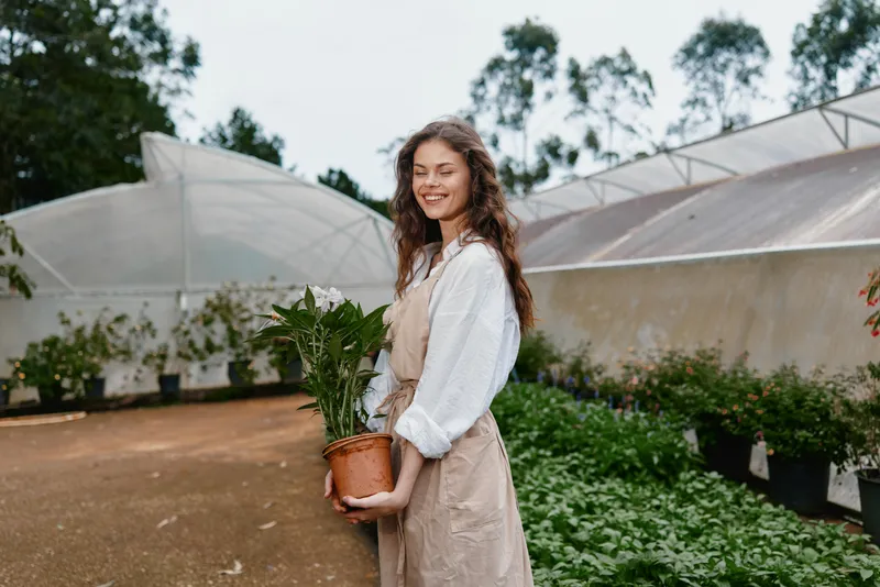 Smiling young woman holding a potted plant in a greenhouse, dressed in casual beige overalls, surrounded by greenery, embodying joy and a connection with nature