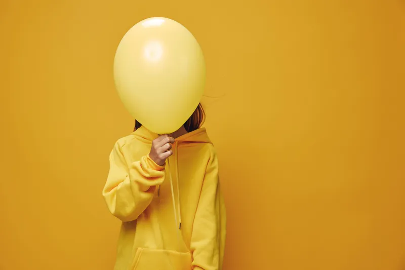 Child in yellow hoodie holding yellow balloon in front of face against yellow background, playful concept with bright colors and casual style in studio portrait.
