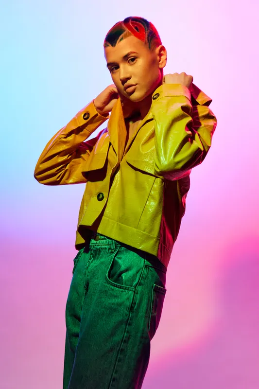 Young woman with short colorful hair wearing a bright yellow jacket and green pants, posing confidently against vibrant blue and pink gradient background.