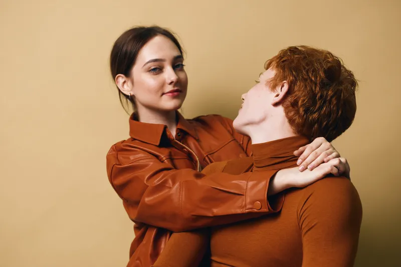 Couple, woman and man dressed in brown clothes hugging and looking at each other with soft smile on beige background. Young people, love, connection and close relationship portrait.