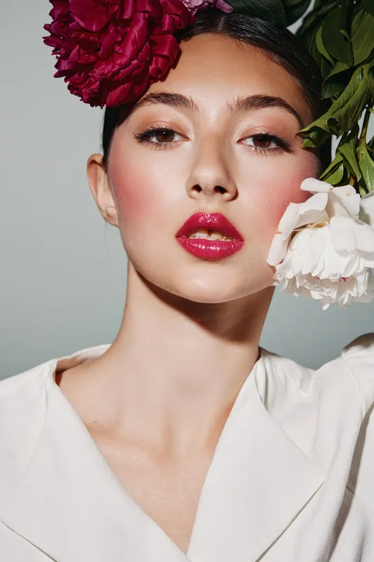 Close-up portrait of a woman with creative bright makeup and bold pink lipstick wearing white holding contrasting colorful flowers. Beauty and fashion concept using artistic styling.