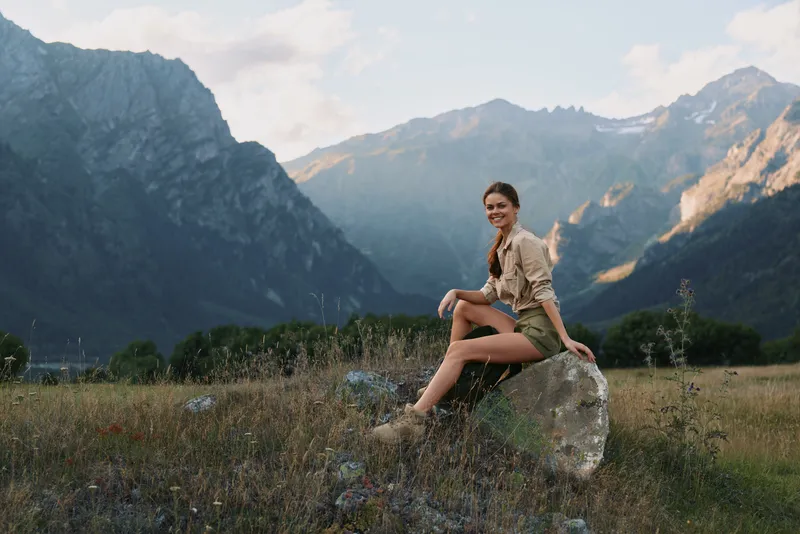 Cheerful woman outdoors, sitting on a rock in a sunlit valley, mountain backdrop, casual outfit, relaxed lifestyle moment, natural colors, expansive landscape