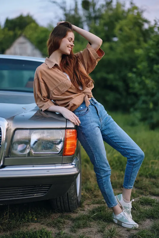 Young woman in casual attire posing by a vintage car, exuding joy and confidence, with a natural background and warm color palette, perfect for lifestyle and fashion themes.