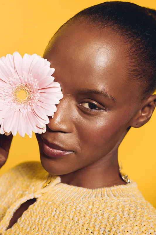 Close-up of a smiling African woman with dark skin holding a pink flower near her face. Yellow background and soft lighting create a warm and cheerful mood. Concept of beauty and nature.