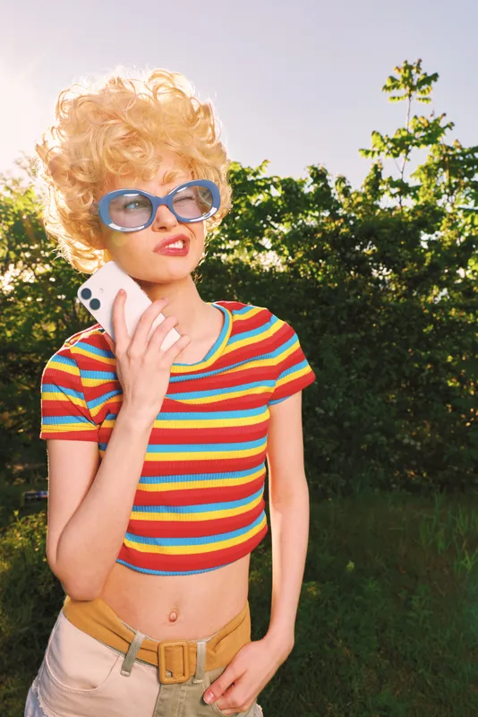 A retro styled woman stands outdoors on a sunny day, striped crop top, blonde wig, blue sunglasses, talking on a mobile. Concept for playful fashion, nostalgia, and carefree lifestyle.