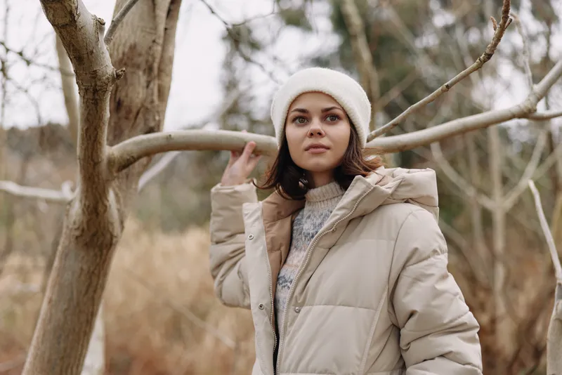 Winter coat portrait of a woman outdoors in a warm jacket and beanie beside bare trees in a calm natural landscape with soft light and calm mood.