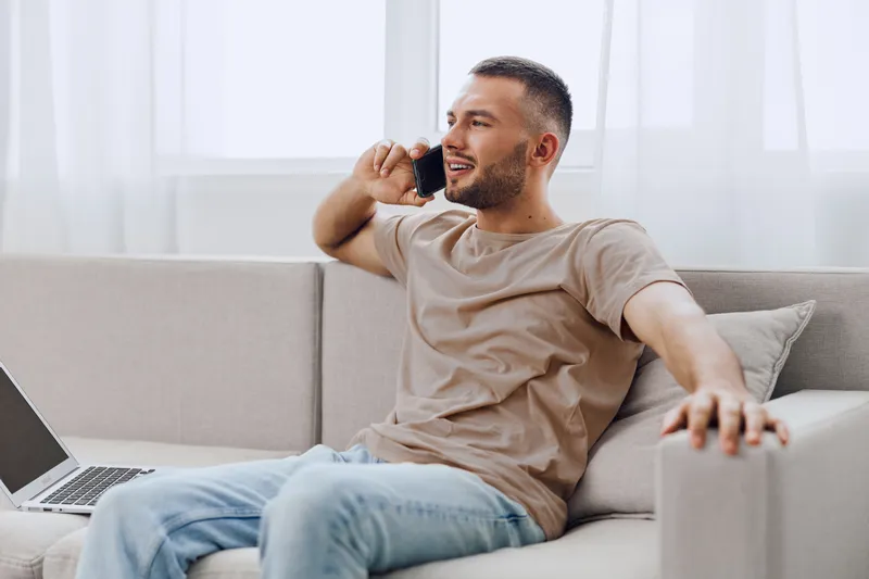 Smiling man engaged in a phone conversation, sitting comfortably on a cozy sofa, dressed casually in a light t-shirt, working at home.