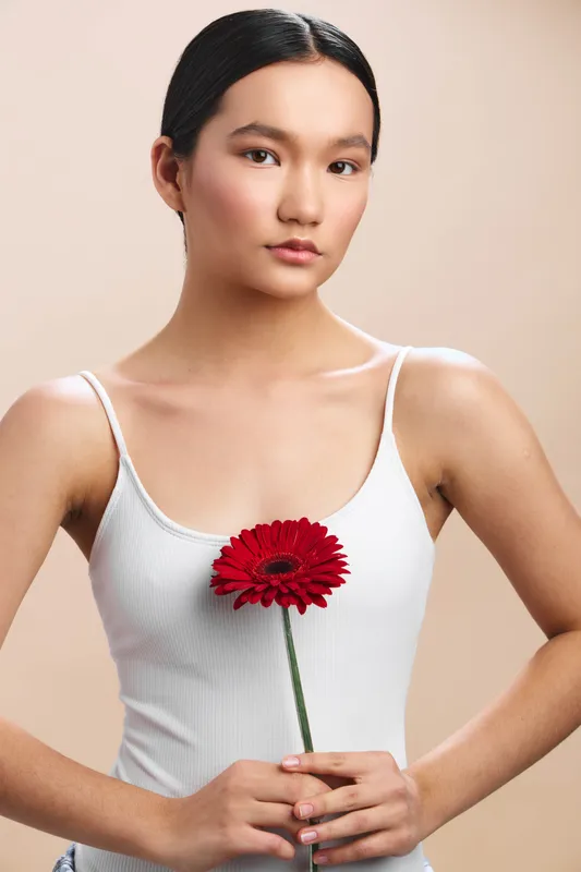 Asian woman wearing a white tank top holds a red flower against a soft neutral background, conveying quiet elegance and simple beauty.
