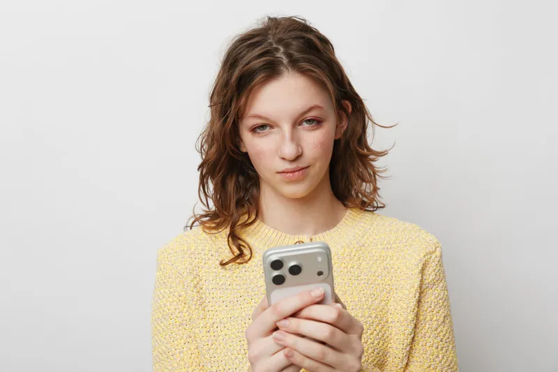 Young woman in yellow sweater looks at smartphone with slight smile, isolated on white background. Concept of communication, technology, social media, and casual lifestyle.