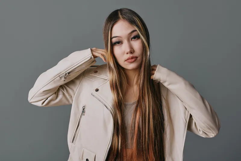 Young woman with long straight hair wearing beige jacket and neutral makeup posing against gray background for fashion portrait and beauty style photography.