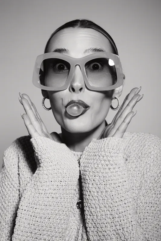 Funny Caucasian woman with pale skin makes playful face and wears large sunglasses and knitted sweater on grey background.