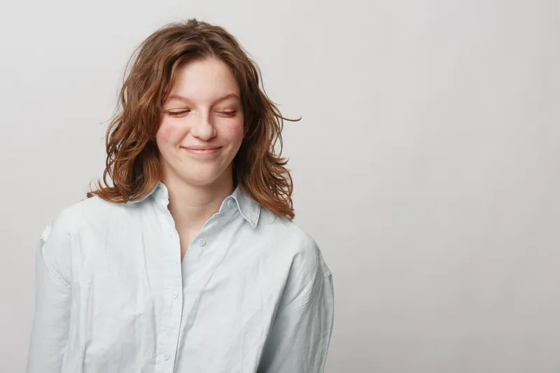 Smiling young woman with curly hair wearing white shirt looks down with closed eyes on light background. Concept of happiness, calmness, and natural beauty in casual style.
