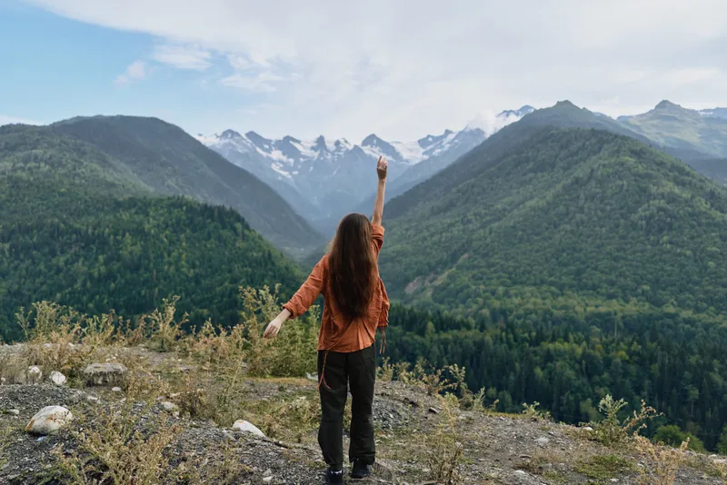 Person stands on a rocky overlook in a warm jacket, raising an arm toward majestic mountains, capturing a moment of outdoor adventure and freedom in nature
