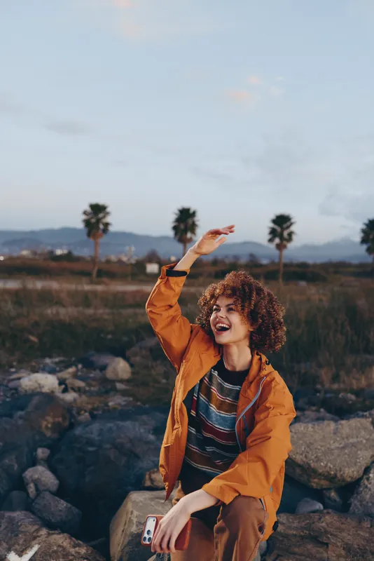 Woman with curly hair in an orange jacket, smiling outdoors on rocks, arms raised in joy, nature landscape with palms and distant hills, lifestyle travel mood, golden sunset light