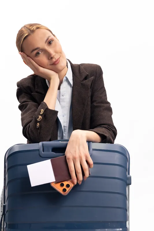 Young woman in smart casual clothes leans on blue suitcase holding passport and phone, isolated on white. Concept of travel, vacation, and business trip preparation.