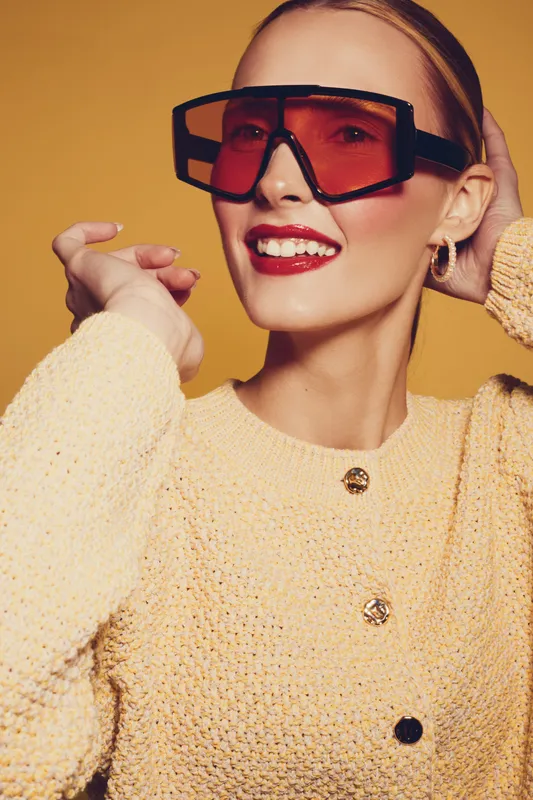 Smiling woman in oversized red sunglasses and yellow knitted sweater poses against orange background. Fashion portrait with bright makeup and trendy accessories for style concept.