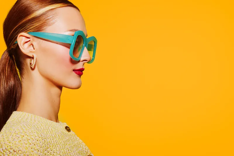 Profile of a stylish woman wearing large turquoise sunglasses and a yellow sweater against a bright orange background. Concept of fashion, summer style, and vibrant colors.