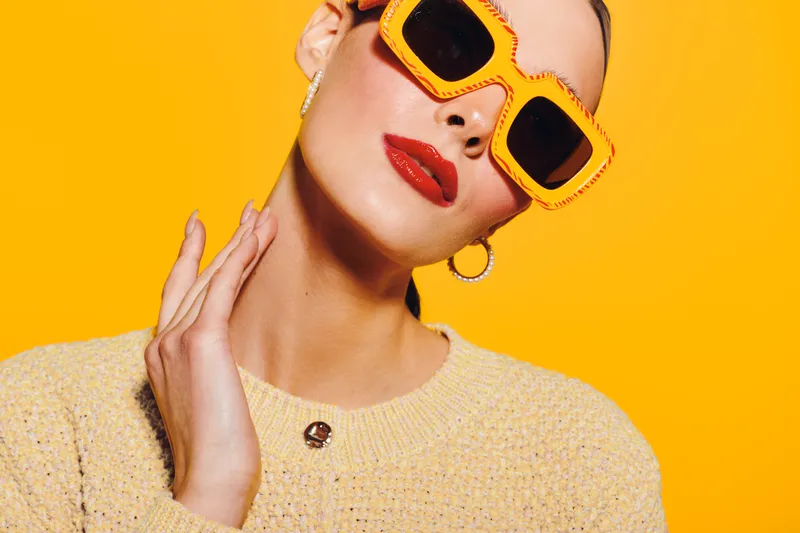 Stylish woman with bright orange sunglasses and red lipstick poses against vibrant yellow background. Fashion portrait with trendy accessories and confident expression.