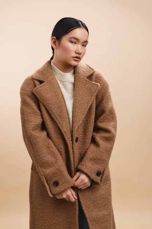 Asian woman wearing a textured brown coat stands against a warm beige backdrop, conveying a calm, stylish mood and confident presence suited for fashion and lifestyle campaigns with a timeless appeal.