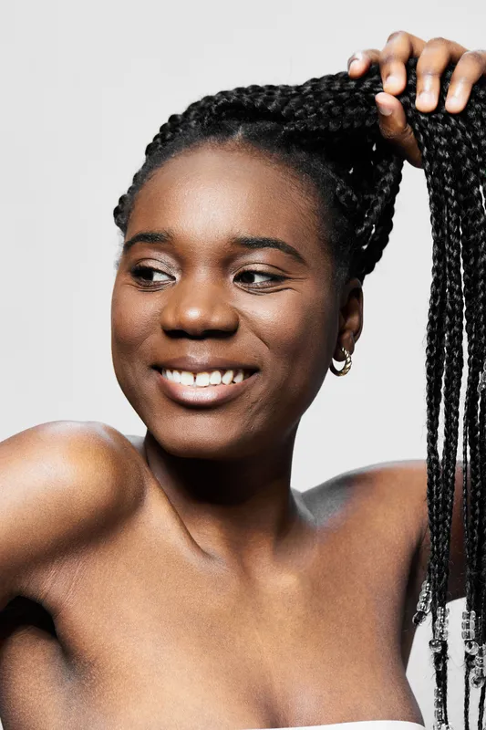 Smiling Black woman with braided hair playfully holding her hair against a neutral background, showcasing confidence and natural beauty