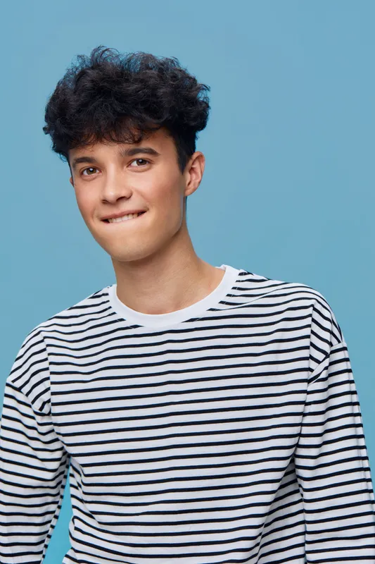 Young man in a striped shirt biting his lip, posing against a light blue seamless background with curly hair and a playful facial expression.