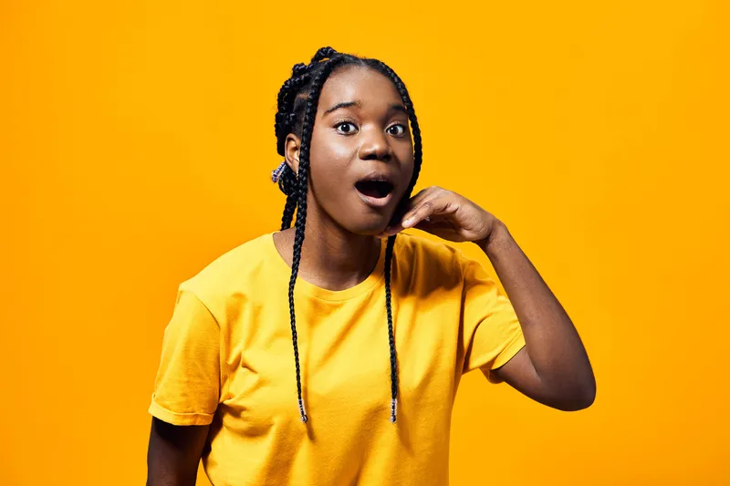Surprised young African woman making a call gesture with her hand, wearing a bright yellow t shirt, set against a vibrant yellow background