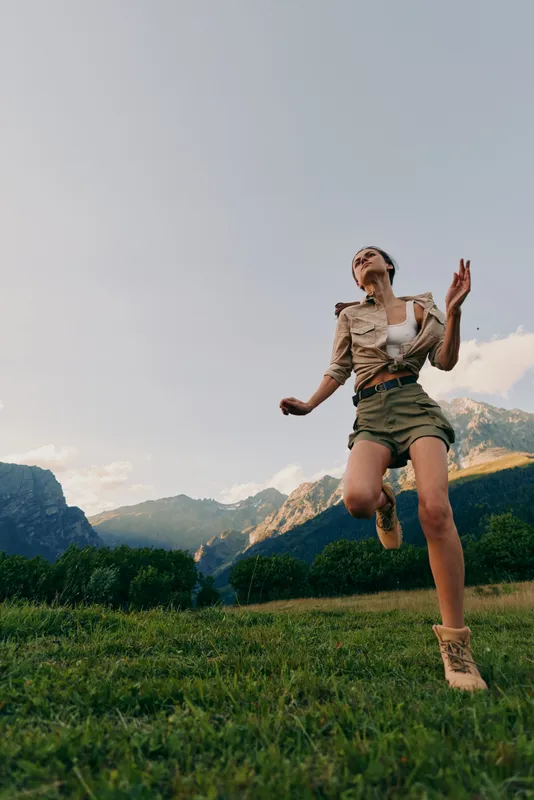 Young woman with short hair jumps joyfully on a grassy field with mountains in the background during a clear day, expressing freedom and happiness in nature.