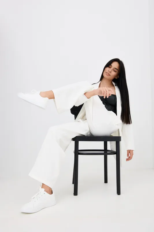 young woman dressed in white suit and sneakers sitting on black chair against white background, long dark hair, relaxed pose, modern minimalism, studio shot