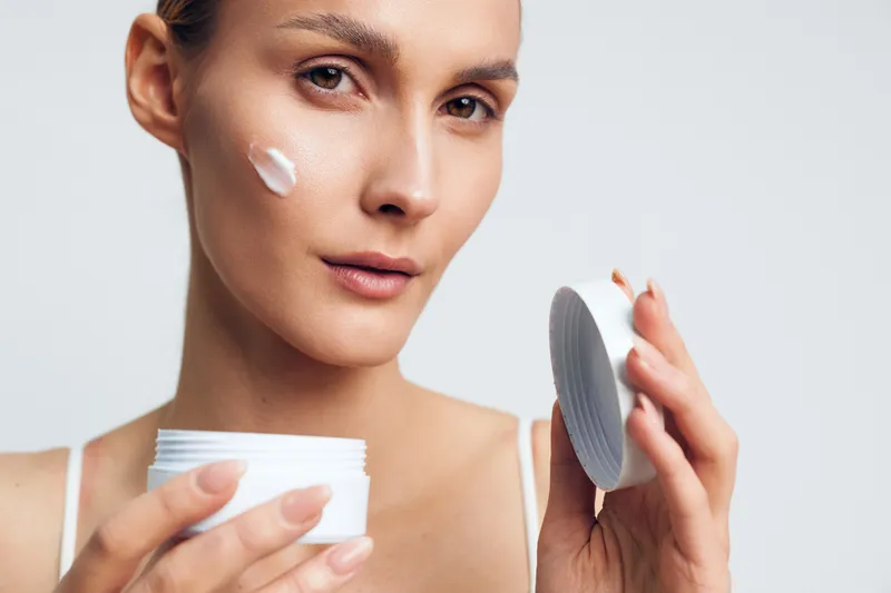 A young woman with clear skin applies face cream from a white jar, holding the lid in one hand against a plain light background. Concept for skincare, beauty, and self-care.
